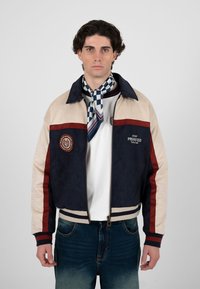 Jeune homme portant une veste zippée bleue, beige et rouge par-dessus une chemise blanche, un foulard bleu à motifs autour du cou, et un jean bleu délavé, faisant face à l'avant.