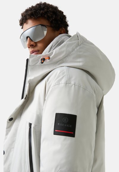 White waterproof jacket with a high collar and large hood, featuring black and red logo patch, zip pockets, and reflective sunglasses.