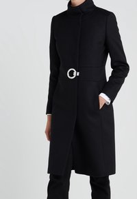 Abrigo negro con cuello alto, mangas largas y una hebilla de cinturón circular plateada. Presenta bolsillos laterales y una silueta recta.