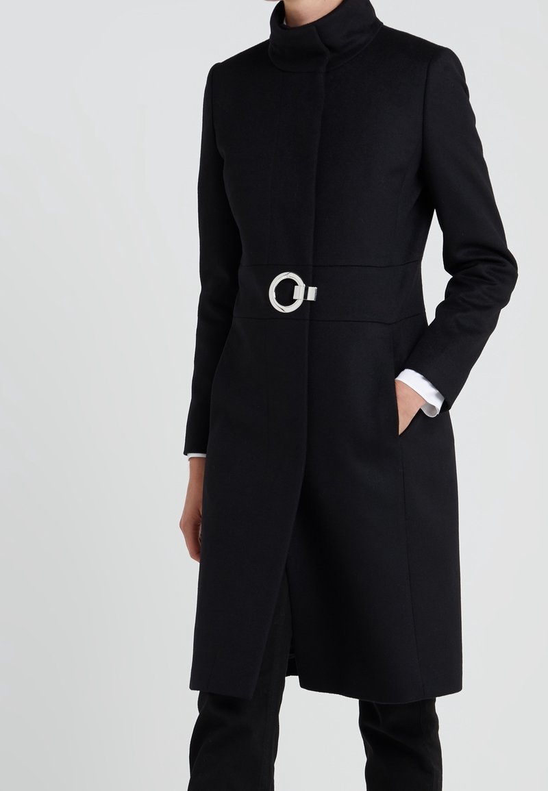 Abrigo negro con cuello alto, mangas largas y una hebilla de cinturón circular plateada. Presenta bolsillos laterales y una silueta recta.
