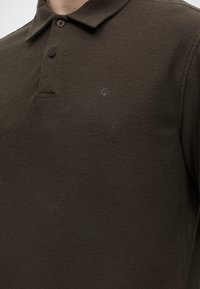 Πολό μπλούζα σε σκούρο ελαιοπράσινο με υφή, με δύο κουμπιά και μικρό στρογγυλό κεντητό λογότυπο κοντά στο στήθος.