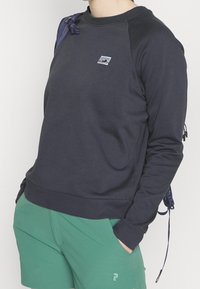 Sudadera marina con mangas raglán, textura suave, que presenta un pequeño gráfico de montaña en el pecho. Combinada con pantalones cortos verdes y una mochila.