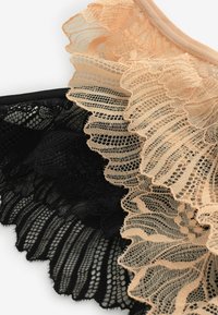 Mežģīņu krūšturis ar viļņotu dizainu, izmantojot ķermeņa krāsas un melnu audumu, izceļot sarežģītas rakstu un faktūru kārtas pretstatītā stilā.