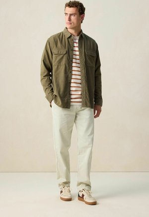 Chemise olive à boutons sur un T-shirt rayé blanc et rouille, associée à un pantalon clair et des baskets aux accents marron.