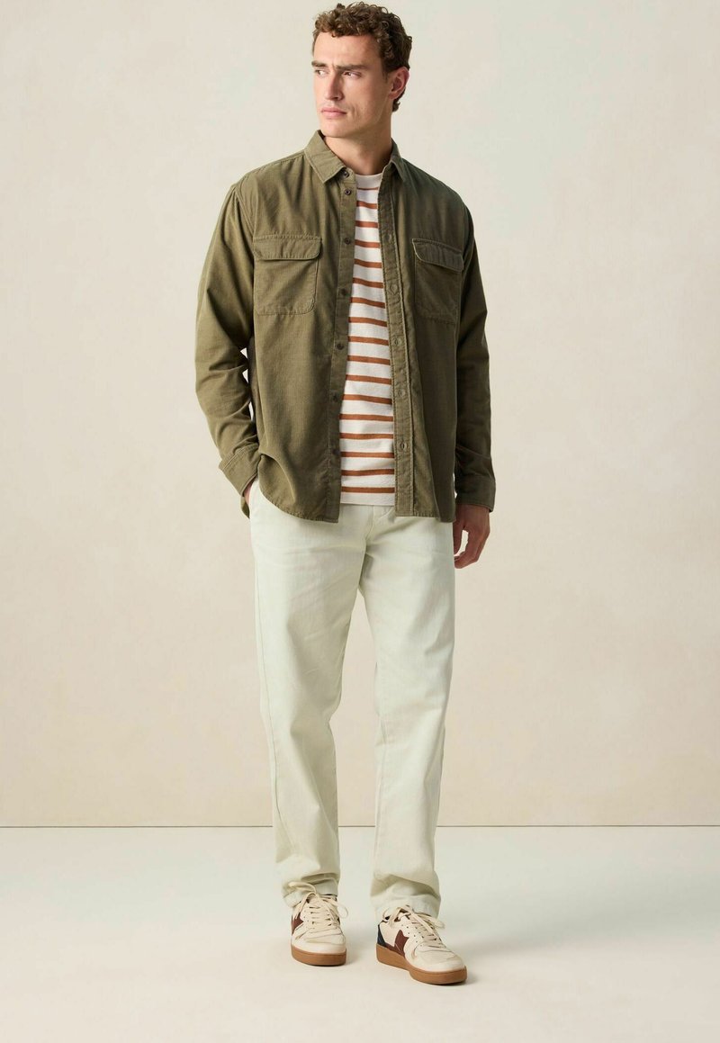 Olijfgroene overhemd met knopen over een wit en roestgestreept T-shirt, gecombineerd met lichte broek en sneakers met bruine accenten.