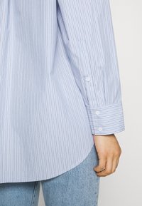 Light blue striped button-up shirt with a soft texture, featuring long sleeves, rounded hem, and white buttons on the cuffs.
