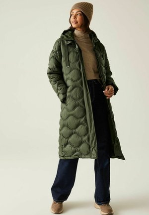 Vrouw die een olijfgroene doorgestikte lange jas draagt, een beige gebreide muts, een beige coltrui, donkere spijkerbroek en beige schoenen.