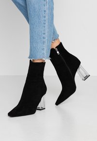 Botines de ante negros hasta el tobillo con punta afilada, que cuentan con un tacón de bloque transparente y una cremallera lateral para un fácil acceso.