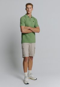 Groene polo met een klassieke kraag en drie knopen, gecombineerd met beige cargoshorts. G gedragen met witte sokken en beige sportschoenen.