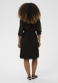 Čierne šaty po kolená s úzkym pásom, priesvitnými rukávmi a splývavou sukňou. Hladká tkanina s jemnou textúrou a ľahkým padaním.