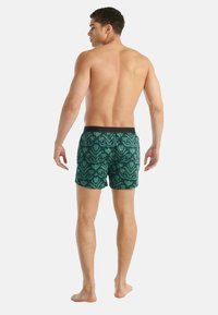 Muž stojící bosý, otočený zády, v zelených boxerkách s květinovým vzorem a černým pasem na bílém pozadí.