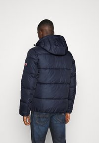 Granatowa kurtka puffer z kapturem, pikowana konstrukcja i wypełnienie. Posiada logo na lewym rękawie i jest połączona z niebieskimi dżinsami.