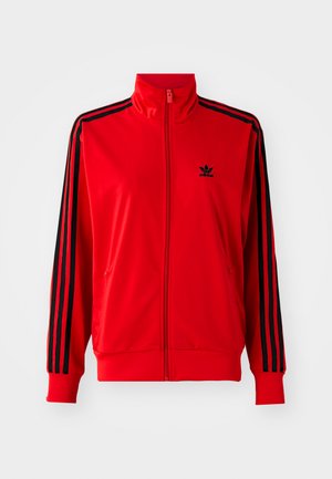 Chaqueta roja con cremallera, hecha de un tejido suave, que presenta rayas laterales negras, un cuello alto y un pequeño logo negro en el pecho.