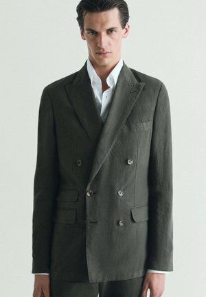 Hombre con el cabello oscuro peinado hacia atrás, vistiendo una chaqueta de traje cruzada de color verde oscuro sobre una camisa blanca con cuello, frente a un fondo sencillo.