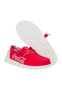 Sneakers di tela rossa con suole in gomma bianca, dotate di logo Coca-Cola e lacci a strisce. La suola presenta un motivo maculato.
