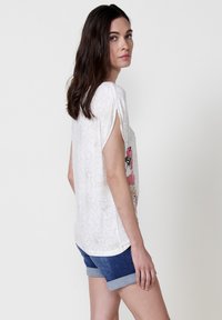 Haut blanc à manches courtes avec un imprimé floral sur le côté, ourlet arrondi et texture douce. Associé à un short en denim avec des ourlets retroussés.