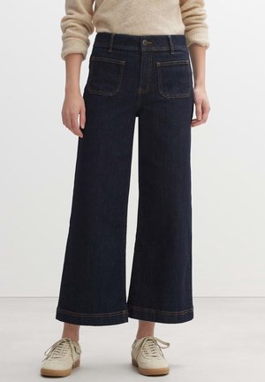 Person wearing dark blue wide-leg cropped jeans with front patch pockets, beige rolled-up sweater, and off-white sneakers.