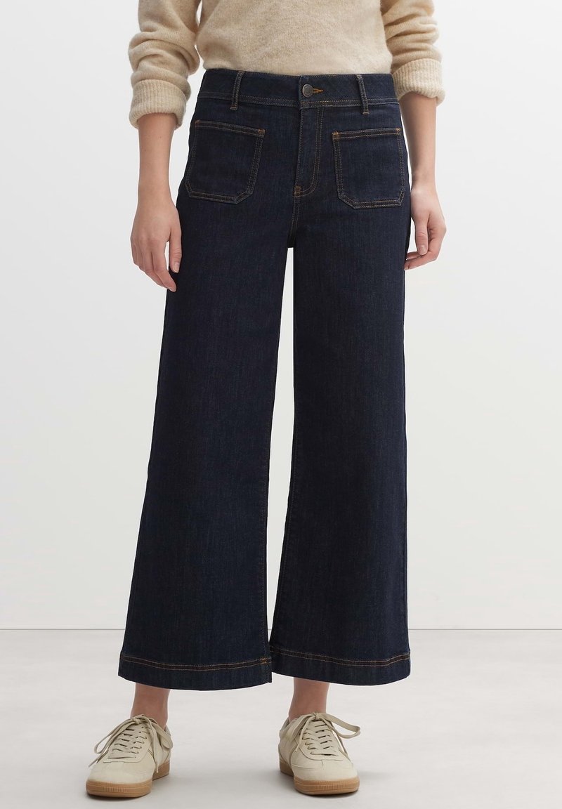 Personne portant un jean large et raccourci bleu foncé avec des poches plaquées à l'avant, un pull beige retroussé et des baskets blanc cassé.
