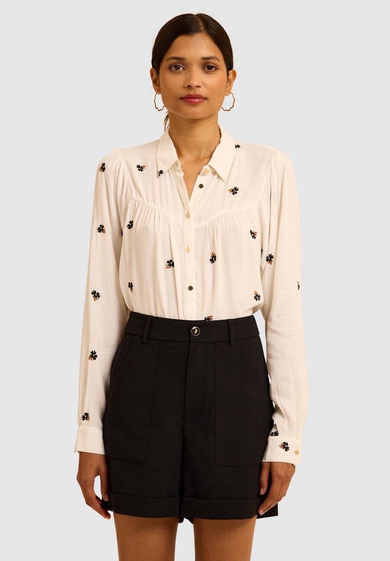 Camicia bianca con ricami floreali neri, maniche lunghe e bottoni, abbinata a pantaloni corti neri a vita alta con tasche frontali.