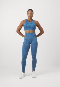 Top curto atlético azul e leggings de cintura alta com um padrão sutil texturizado. Modelo usa ténis brancos, posando diante de um fundo simples.
