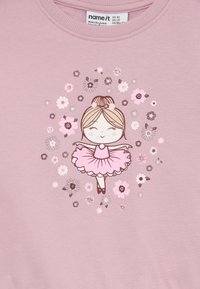 Smilende ballerina i en pink tutu med blomster omkring hende på en lys lilla stofbaggrund, vist på en trøje i børnestørrelse.