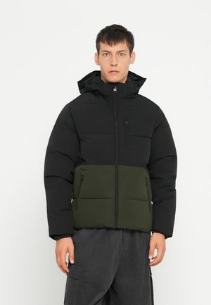 Mladý muž nosi čiernu a tmavozelenú prešívanú bundu s kapucňou a vreckami na zips, stojaci pred jednoduchým bielym pozadím.