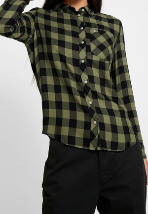 Kvinde iført en olivengrøn og sort ternet skjorte med brystlomme og sorte bukser foran en ensfarvet baggrund.