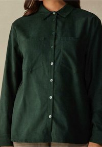 Camicia verde scuro con bottoni realizzata in tessuto morbido, con colletto, due tasche sul petto e bottoni blu chiaro lungo la parte anteriore.