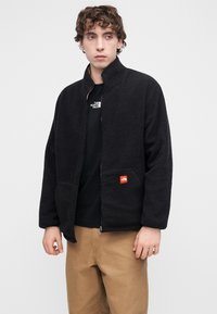 Μαύρο φλις τζάκετ με ψηλό γιακά, κλείσιμο με φερμουάρ και ένθετο πορτοκαλί λογότυπο σε αντίθεση. Φορώντας το πάνω από μαύρο μπλουζάκι, συνδυασμένο με χακί παντελόνι.