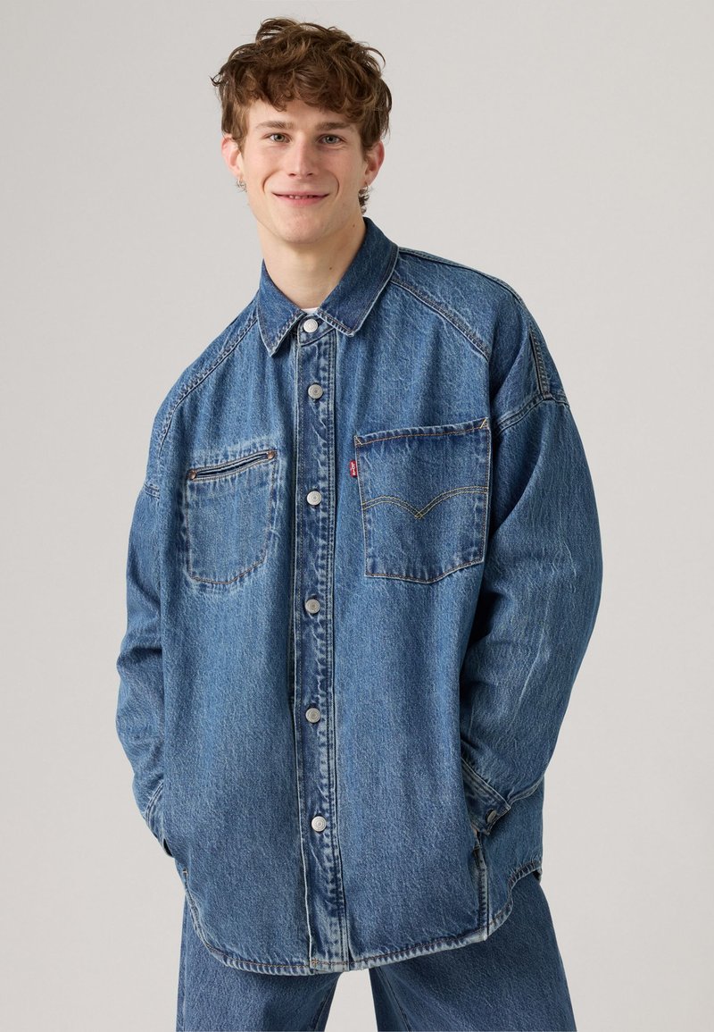 Lys blå denimskjorte med knapplukking i front, spiss krage og to brystlommer. Har søm detaljer og en løs passform.