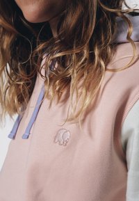 Světle růžová mikina s kapucí v levandulové barvě, s malým vyšitým logem slona a texturovaným materiálem, který představuje neformální design.