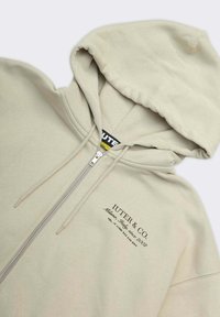 Felpa con zip di colore beige chiaro in morbido misto cotone. Caratterizzata da un cappuccio con cordino, zip argento e logo stampato in nero sul davanti.