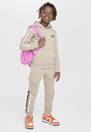 Felpa beige con logo Nike nero, pantaloni cargo beige abbinati con tasche laterali e sneakers arancioni e bianche. Zaino rosa con dettagli in rete.