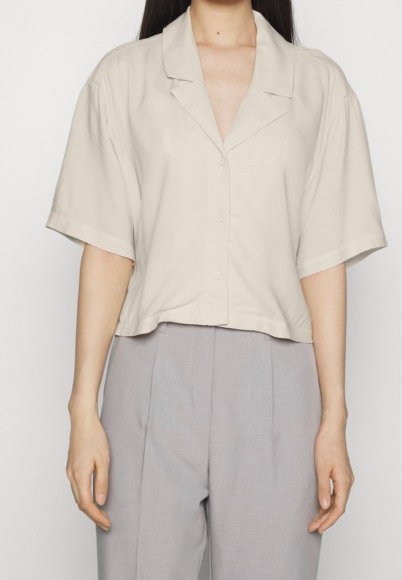 Femme portant une chemise ample beige à manches courtes avec boutons et un pantalon ample taille haute gris clair, sur un fond uni.