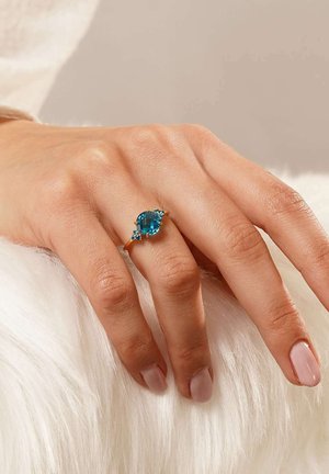 Goldring mit einem großen blauen Edelstein, flankiert von kleineren blauen Akzenten, auf einem strukturierten weißen Furr Hintergrund.