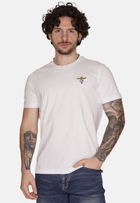 T-shirt in cotone bianco a maniche corte con un piccolo distintivo ricamato in oro e rosso sull'area del petto sinistro. Design semplice, girocollo.
