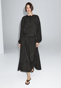 Langes schwarzes Kleid mit lockerer Passform, gerafftem Ausschnitt, Ballonärmeln und einem fließenden Rock. Kombiniert mit schwarzen Flip-Flops. Glatte Textur.