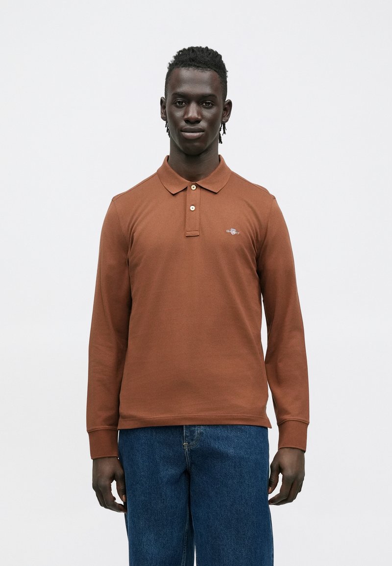 Poloshirt met lange mouwen in bruin katoen met een kraag, twee knopen en een klein logo op de borst. Draag het met blauwe spijkerbroek.