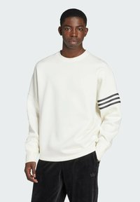 Sweatshirt blanc avec un col rond, des épaules tombantes et trois rayures noires sur la manche gauche. Associé à un pantalon en velours noir.