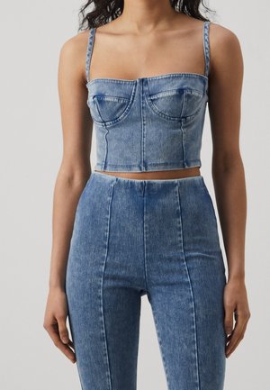 Vrouw draagt een lichtblauwe crop top van denim met dunne bandjes en een high-waisted skinny jeans, staand tegen een effen achtergrond.