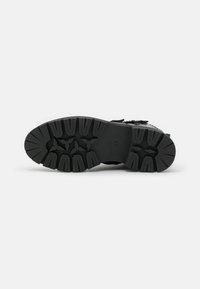 Semelle de chaussure de sport noire avec des crampons profonds et des motifs géométriques, composée d'un matériau en caoutchouc résistant et d'un talon renforcé pour une meilleure adhérence et stabilité.