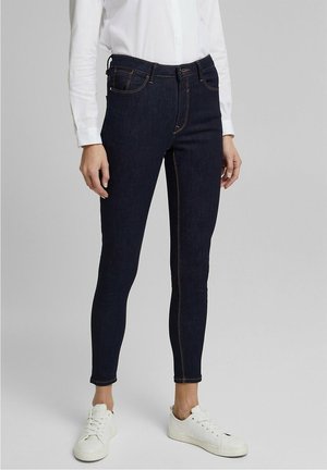 Osoba ubrana w ciemne obcisłe dżinsy, białą koszulę z długim rękawem i białe trampki, stojąca na jednolitym jasnym tle.