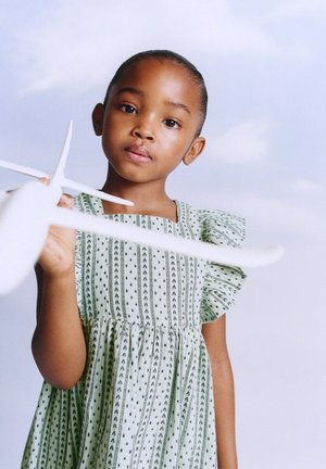 Jong meisje in een jurk met patroon, houdt een wit modelvliegtuig vast en kijkt ernaar tegen een achtergrond van een lichtblauwe lucht.