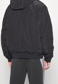Veste noire à capuche avec une texture froissée, des poignets et un ourlet élastiques, au coupe décontractée. Portée par-dessus un pantalon gris foncé avec des rayures sur les côtés.
