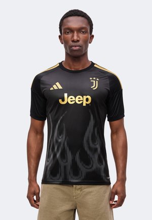 Homme portant un maillot de football noir de la Juventus avec des logos dorés et un motif de flammes, associé à un pantalon beige sur fond uni.