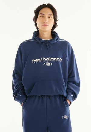 Junge Person trägt einen dunkelblauen New Balance Hoodie und passende Jogginghose mit den Händen in den Taschen, stehend vor einem schlichten hellen Hintergrund.