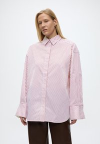 Ξανθιά γυναίκα που φοράει υπερμεγέθη λευκό πουκάμισο με κόκκινες κάθετες ρίγες και φαρδιά μανσέτα, συνδυασμένο με σκούρο καφέ παντελόνι, στέκεται μπροστά σε λευκό φόντο.