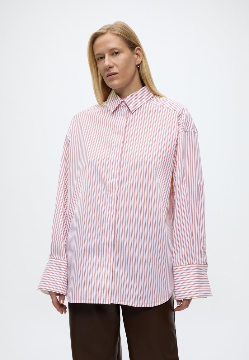 Ξανθιά γυναίκα που φοράει υπερμεγέθη λευκό πουκάμισο με κόκκινες κάθετες ρίγες και φαρδιά μανσέτα, συνδυασμένο με σκούρο καφέ παντελόνι, στέκεται μπροστά σε λευκό φόντο.