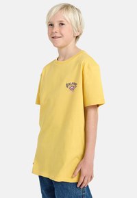 Geel katoenen t-shirt met korte mouwen en een ronde halslijn, met een paarse grafische print op de linkerkant van de borst. Gekocht met blauwe spijkerbroeken.