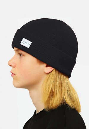 Black ribbed beanie with a folded cuff, featuring a white label stitched near the front. The model has long blonde hair.